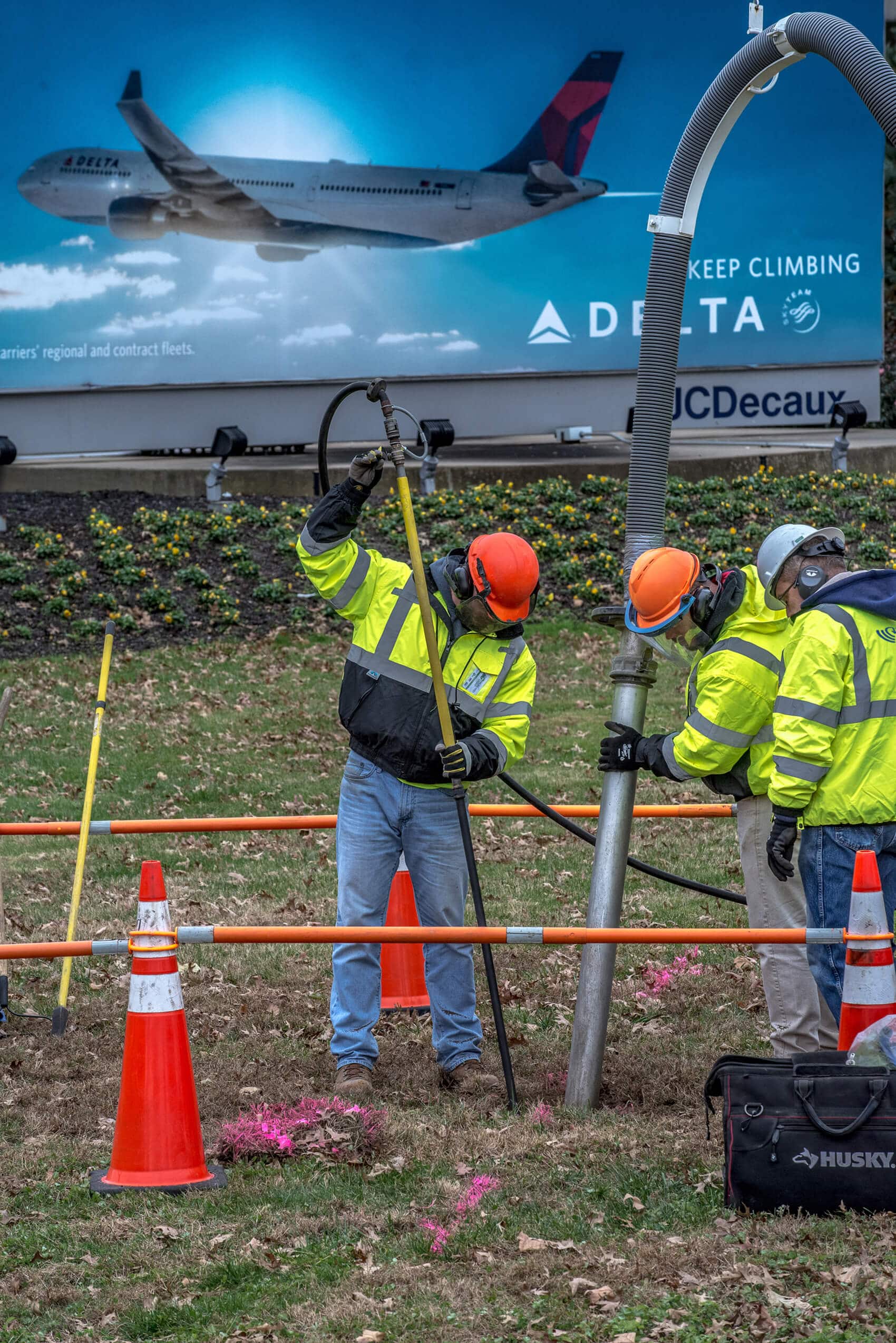 Three men using an air lance and suction to excavate a grassy area with a billboard in the background