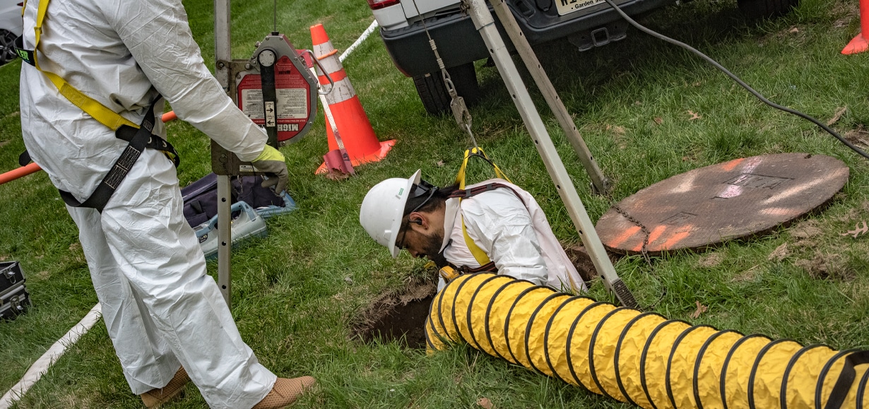 Man in construction safety equipment entering a manhole in a grassy area.