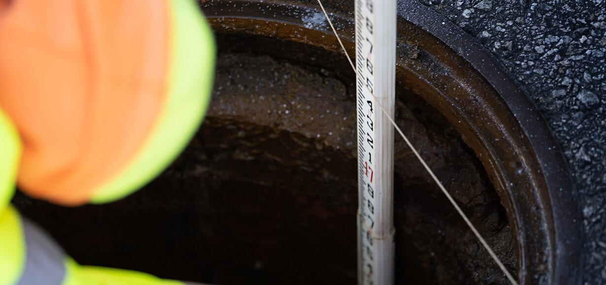 Man wearing neon safety gear measuring a manhole's depth using a long measuring pole.